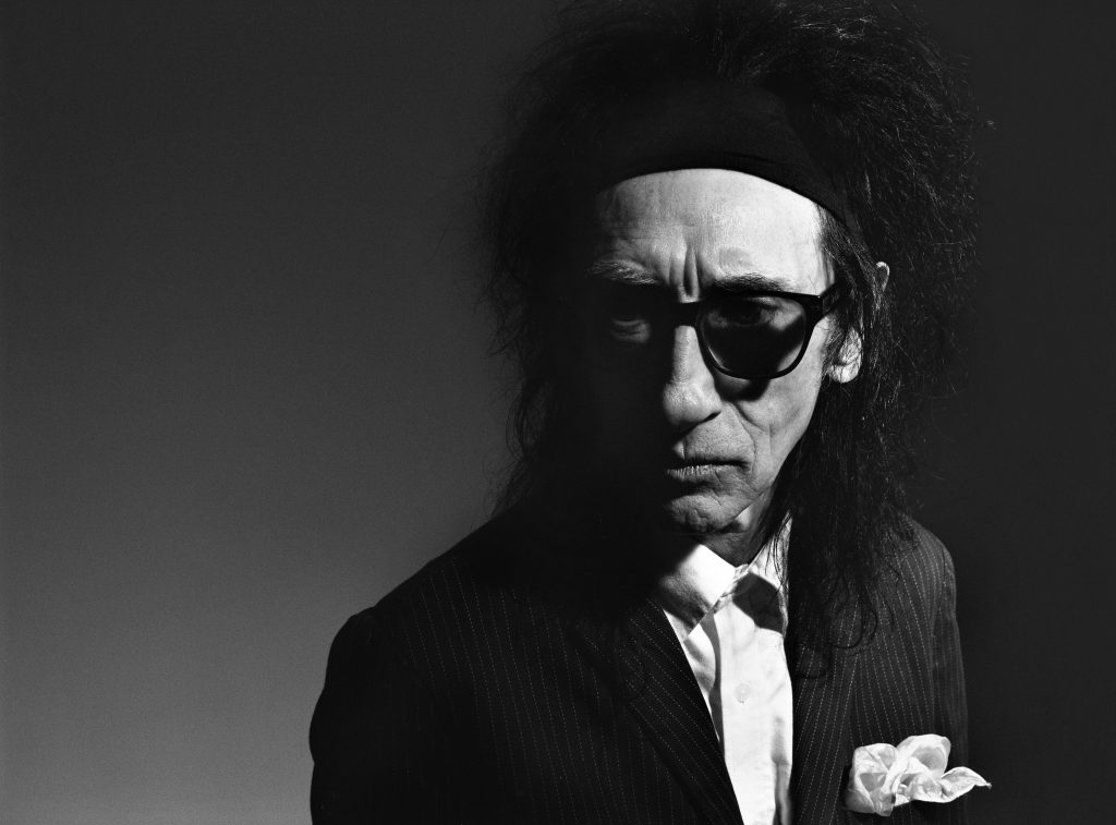 John Cooper Clarke stands to the right side of the picture, a white handkerchief poking out of his suit jacket pocket. He is wearing dark rimmed glasses and has jet black messy hair. I Wanna Be Yours