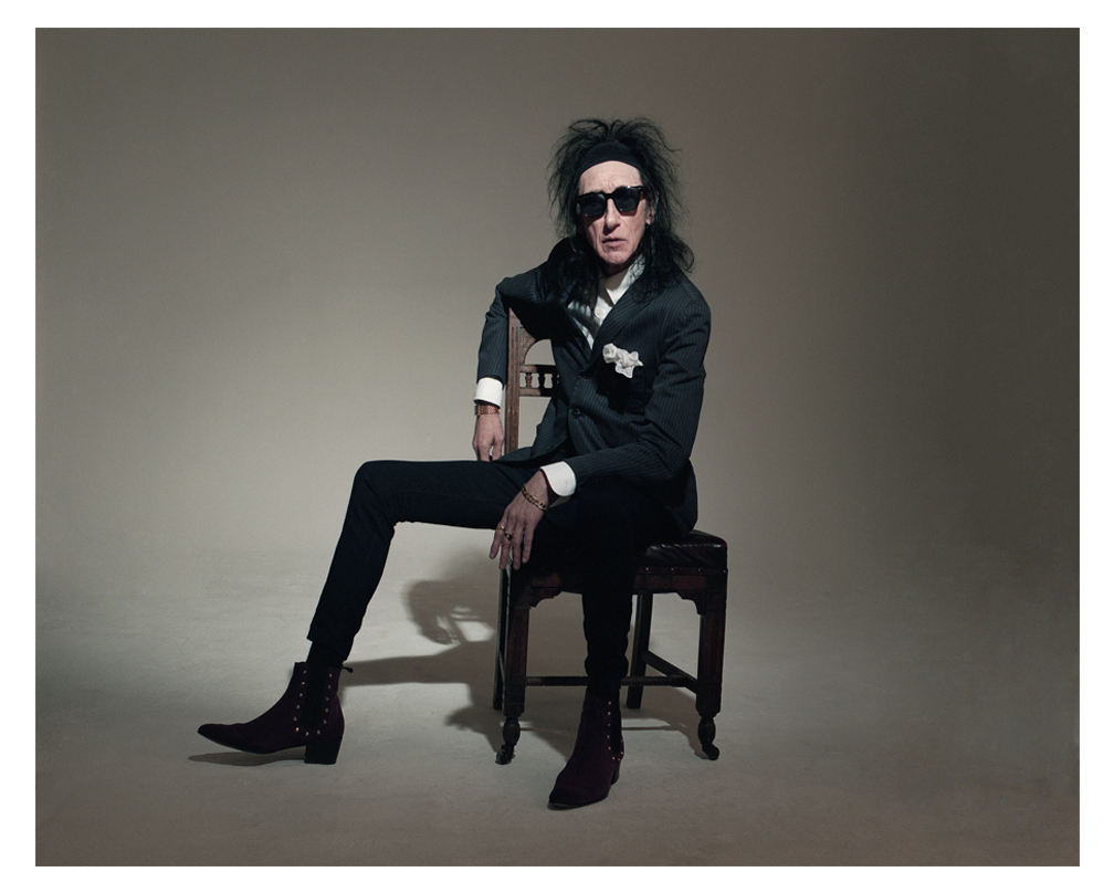 John Cooper Clarke sits in an old wooden chair, dressed in his trademark black suit, white shirt, along with dark rimmed glasses and his dark messed up hair leading to one of his many nicknames, Johnny Clarke, the name behind the hairstyle.