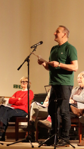 Christopher Eccleston performing poetry on stage into a microphone.