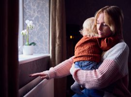 Mother With Son Trying To Keep Warm By Radiator At Home During Cost Of Living Energy Crisis